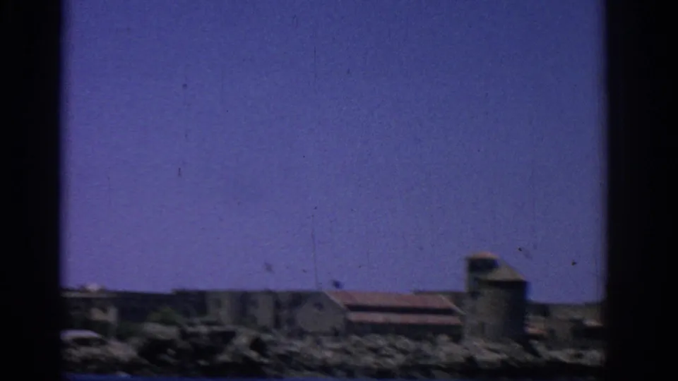 1962 Rhodes shoreline view with a small stone tower-like structure, red roof, and coastal buildings.