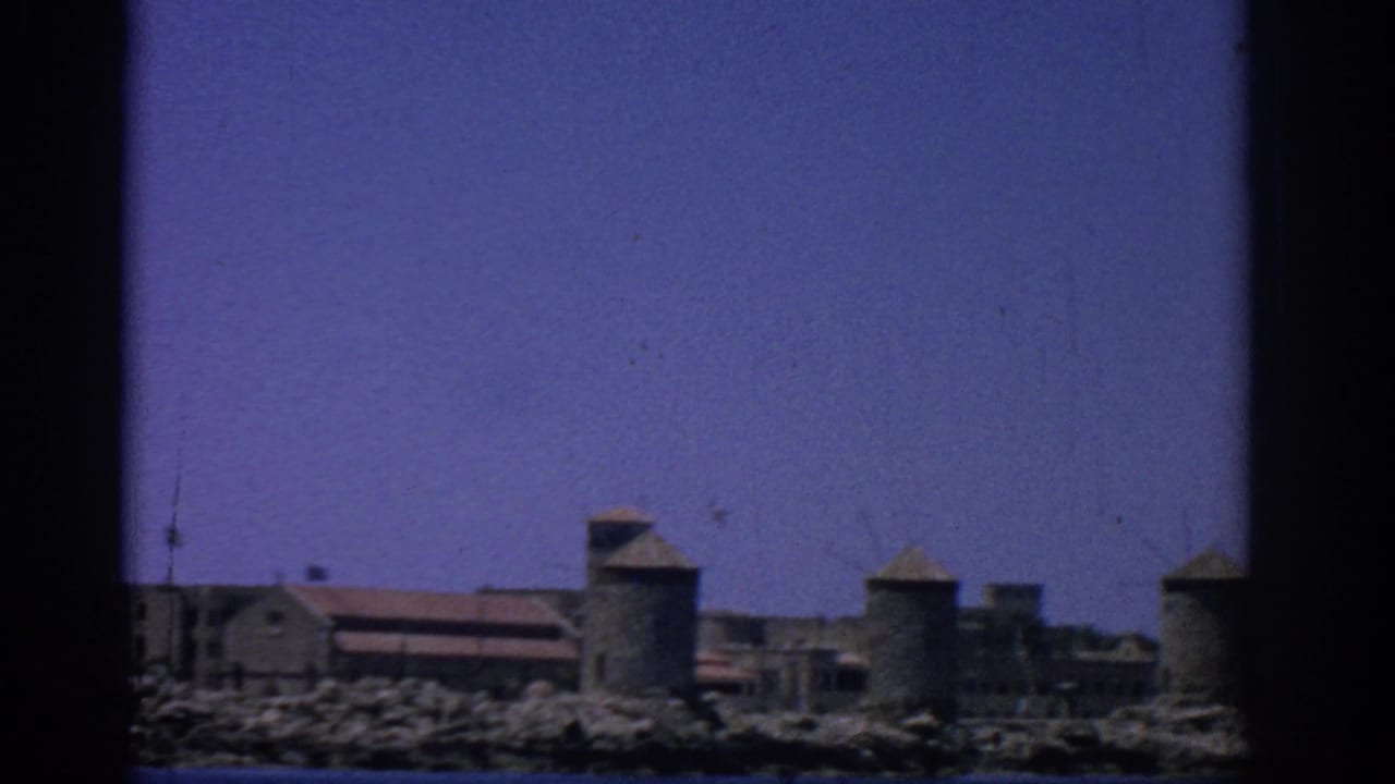 Coastal stone buildings and round towers seen across blue water under a clear sky.