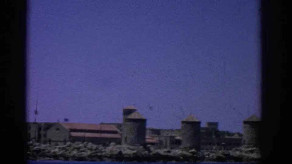 1962 8mm view of Rhodes, Greece, showing a rocky waterfront with stone buildings and cylindrical towers across the sea.
