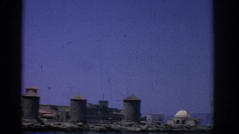 Rhodes, Greece waterfront in 1962 with stone towers, rocky shore, blue sea, and a domed building at the right edge.