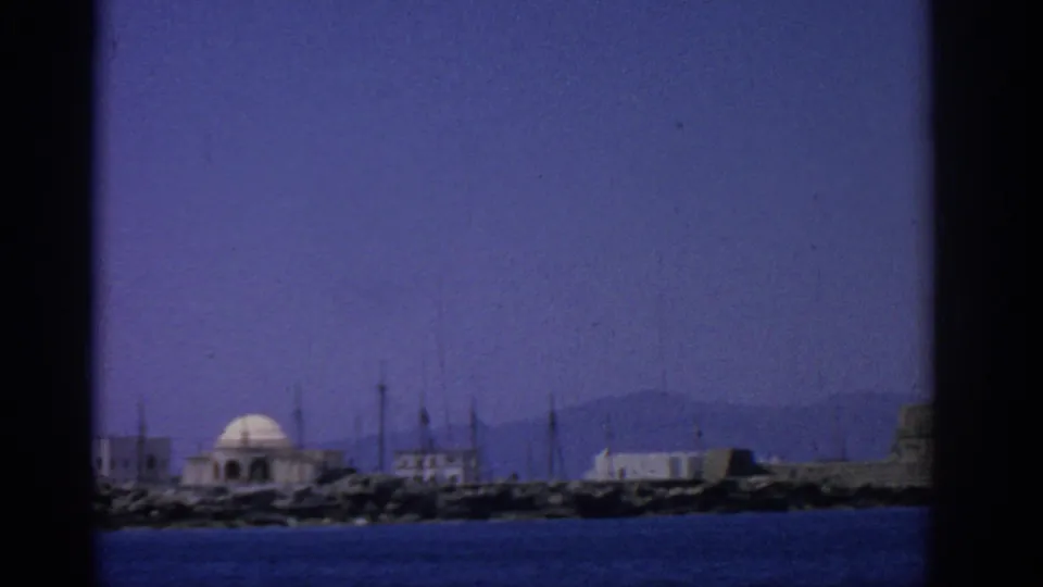 1962 coastal view in Rhodes, Greece, with a domed building, low waterfront structures, masts, distant hills, and blue water.
