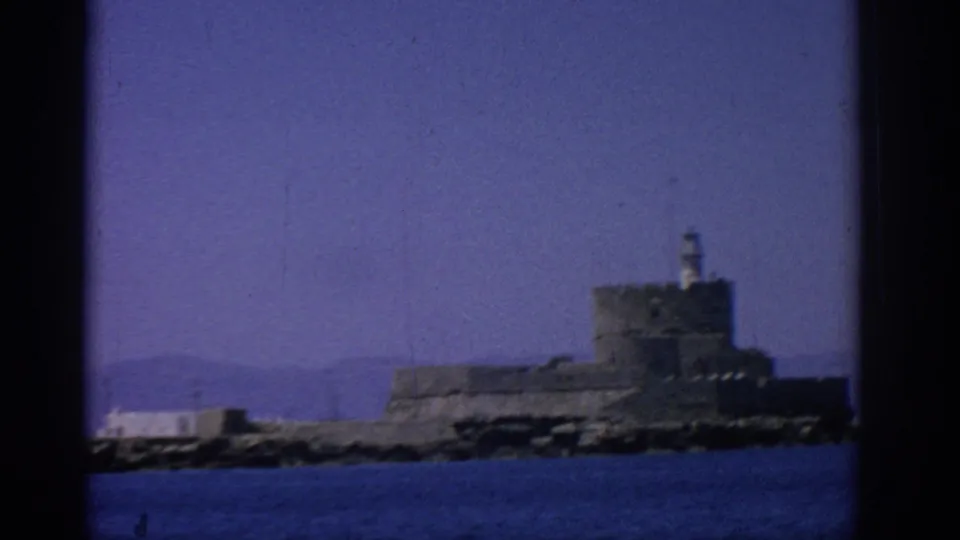 Rhodes, Greece 8mm frame showing a fortified waterfront structure with a small lighthouse-like tower, distant hills, and sea foreground.