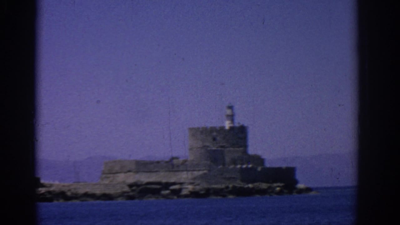 Distant stone fortress with a small lighthouse across blue sea water.
