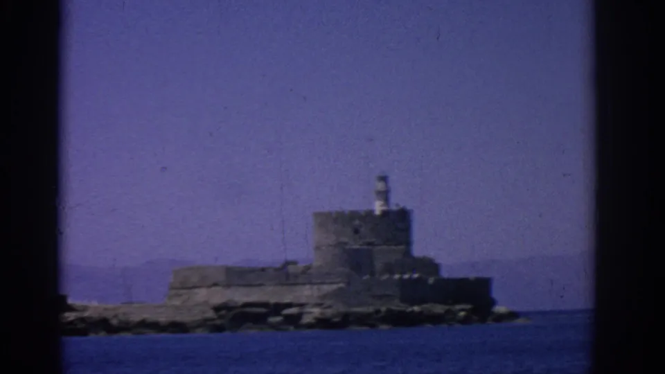 1962 Rhodes home-movie frame showing a distant grey stone sea fortress and small lighthouse beyond blue water.