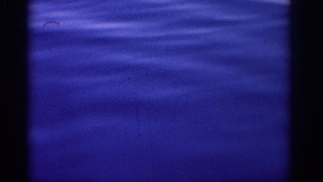 Blue-tinted 8mm view of the sea near Rhodes, with faint ripples, soft focus, and small physical-film marks.