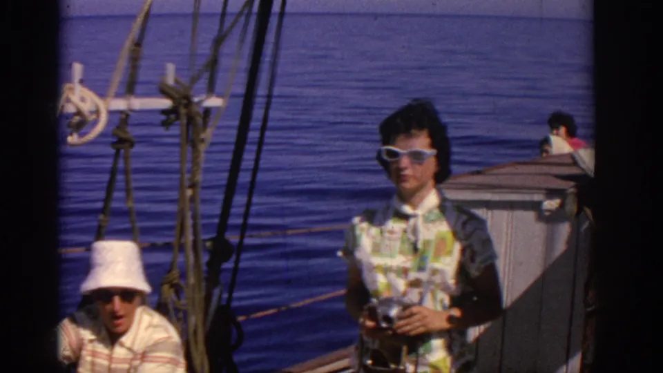 1962 Rhodes home-movie frame showing a woman in sunglasses holding a camera and a seated man in a white hat on a sailboat deck.