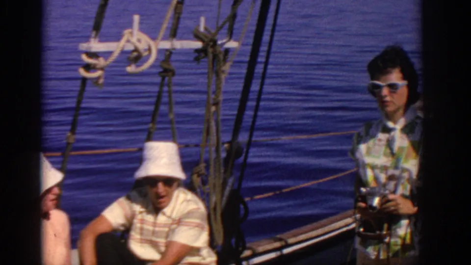 1962 Rhodes sailing frame with a woman holding a camera, a seated man in a white bucket hat, boat rigging, and calm blue sea.