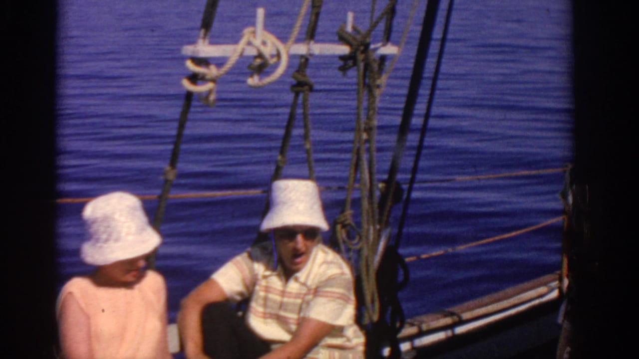 Two adults in white hats sit on a sailboat with blue water and rigging behind them.