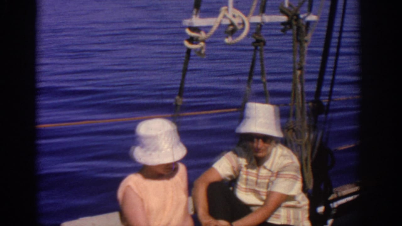 A woman and man in hats sit close together on a sailboat with calm blue water behind them.