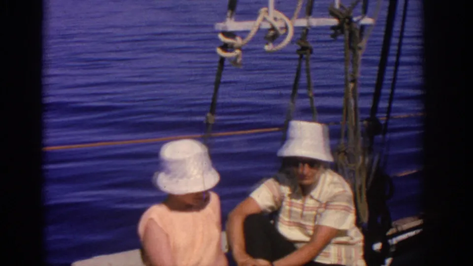 The couple remains seated on the sailboat in Rhodes, Greece, wearing white hats and sunglasses against a blue sea backdrop.