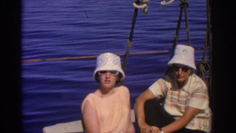 A 1962 home-movie frame shows the couple looking toward the camera on a sailboat in Rhodes, with rigging and rippling sea behind them.