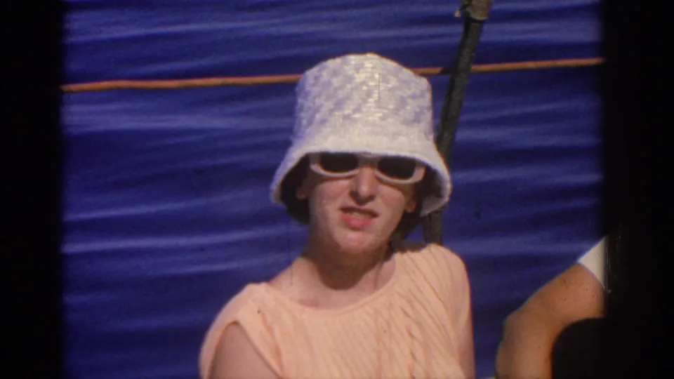 A close 1962 Rhodes sailing shot shows the woman in a white textured hat, sunglasses, and peach dress against blue water.