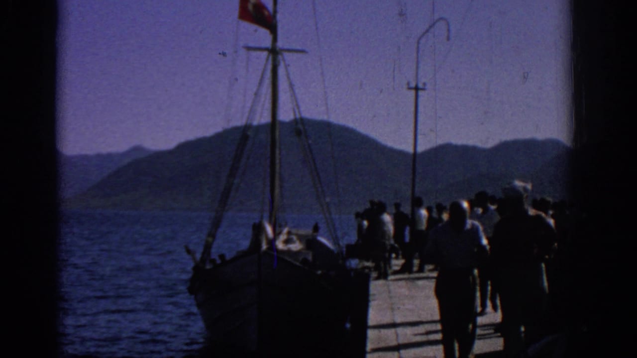 1962 Rhodes home-movie frame showing a pier crowd near a wooden boat with a Turkish flag against blue water and distant hills.