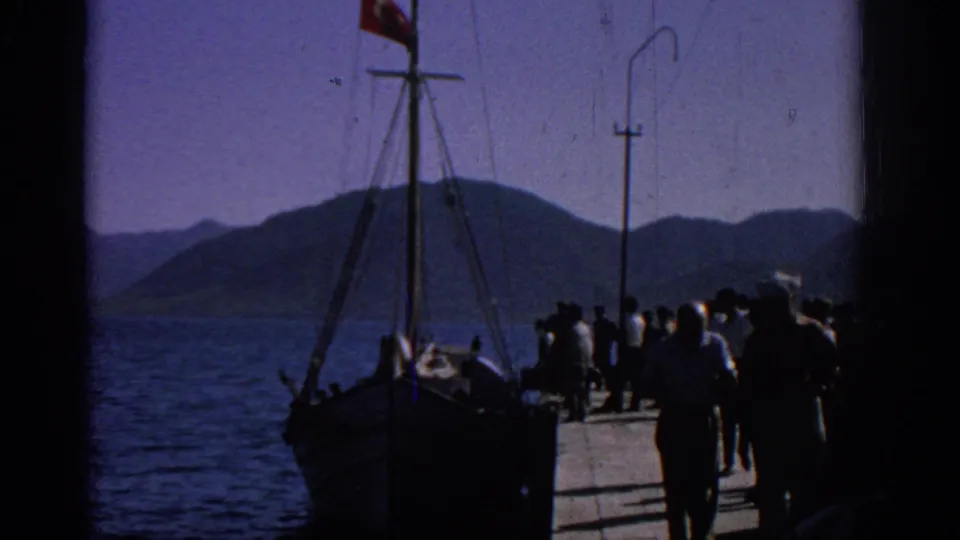 1962 Rhodes home-movie frame showing a pier crowd near a wooden boat with a Turkish flag against blue water and distant hills.