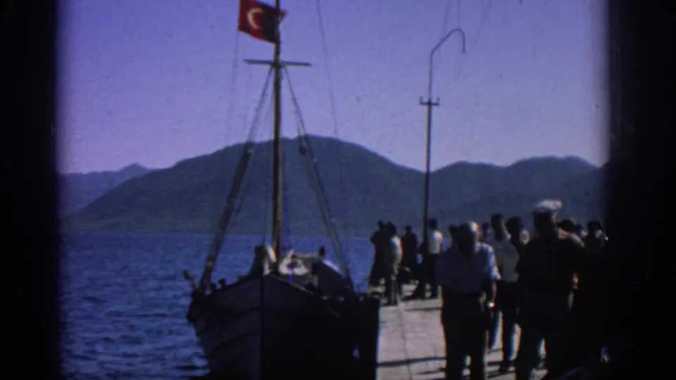 Rhodes, 1962 footage frame with people on a pier next to a moored wooden boat carrying a Turkish flag.