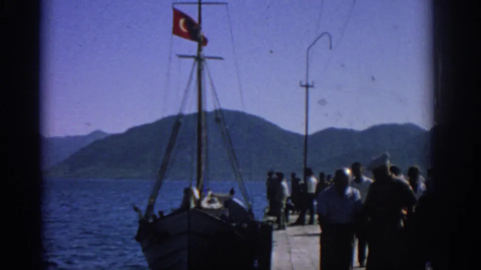 1962 Rhodes pier scene with visitors in 1960s clothing near a Turkish-flagged wooden boat and mountainous shoreline.