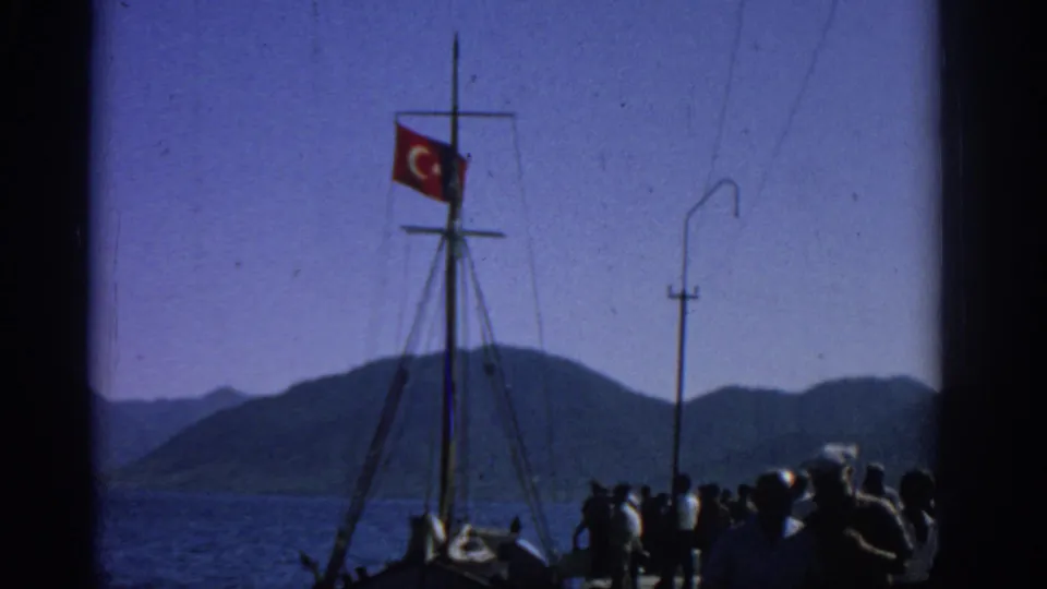 Home-movie frame from Rhodes in 1962 showing a Turkish flag on a boat mast above people gathered on a waterfront pier.