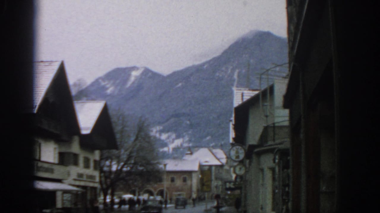 1963 8mm view of a snowy European town street near Munich, with rooftops, distant pedestrians, and mountains beyond.
