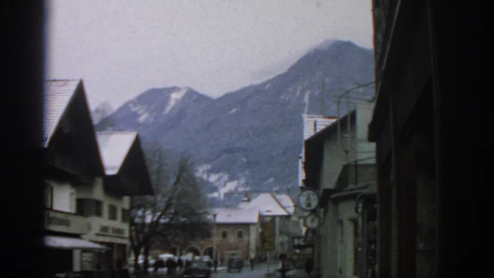 1963 8mm view of a snowy European town street near Munich, with rooftops, distant pedestrians, and mountains beyond.