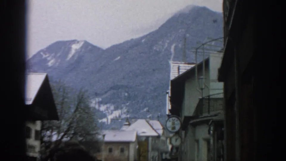 1963 home-movie frame near Munich showing a winter street, snow on roofs, bare trees, and mountains behind the buildings.