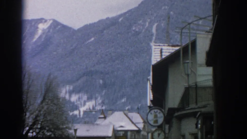 1963 8mm frame near Munich with a closer look at snow-covered rooftops, a round clock sign, winter trees, and mountain slopes.