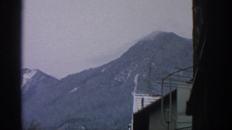 1963 home-movie view near Munich showing a mountain slope with snow patches above town buildings and a partially visible clock sign.