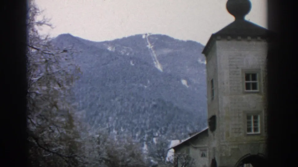 1963 Munich home-movie frame showing a pale tower, arched passage, bare trees, rooftops, and snow-dusted mountains.