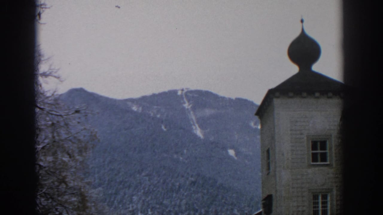 1963 Munich-area 8mm frame showing a snow-dusted wooded mountain behind an onion-dome building, partly blocked by dark foreground edges.