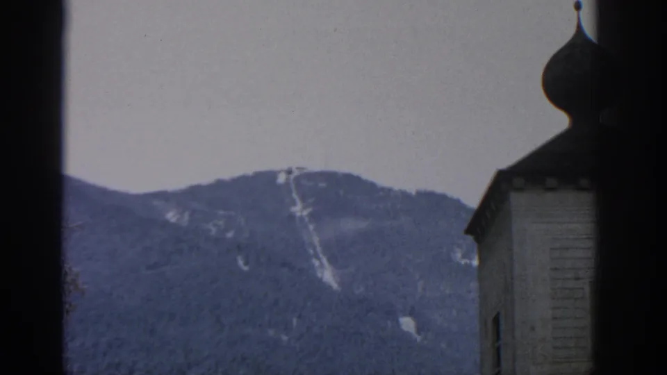 1963 Munich-area 8mm view of a wooded mountain ridge with pale snow traces and an onion-dome structure partly visible on the right.