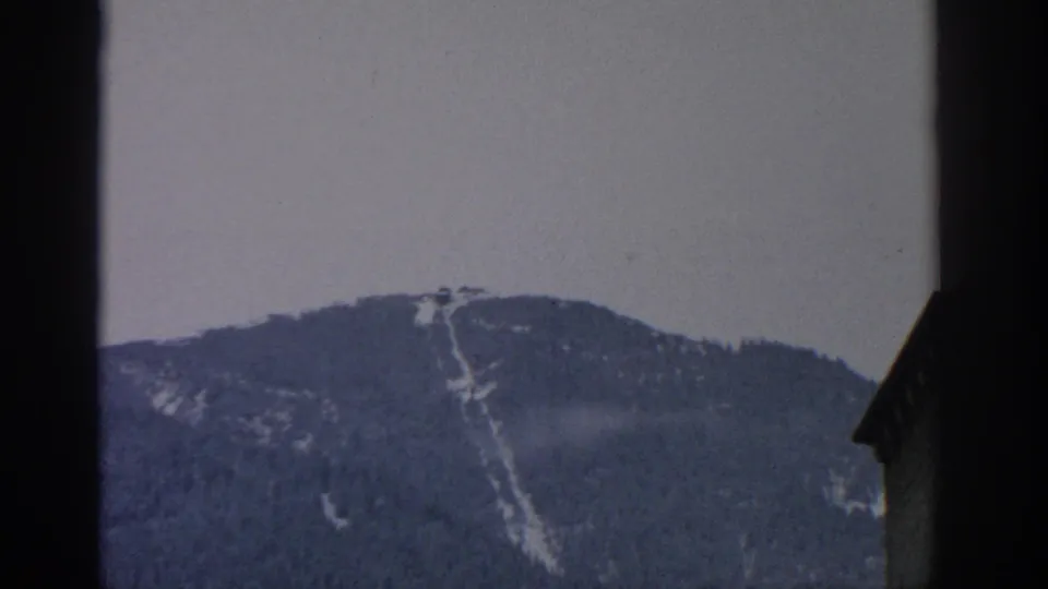 1963 Munich-area 8mm frame centered on a tree-covered mountain slope crossed by a pale vertical run, with a dark building edge in the foreground.