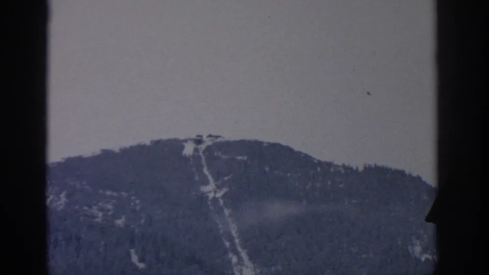 1963 Munich-area 8mm view of a wooded mountain crest with a pale snow or ski run descending the slope, partly framed by a dark right edge.