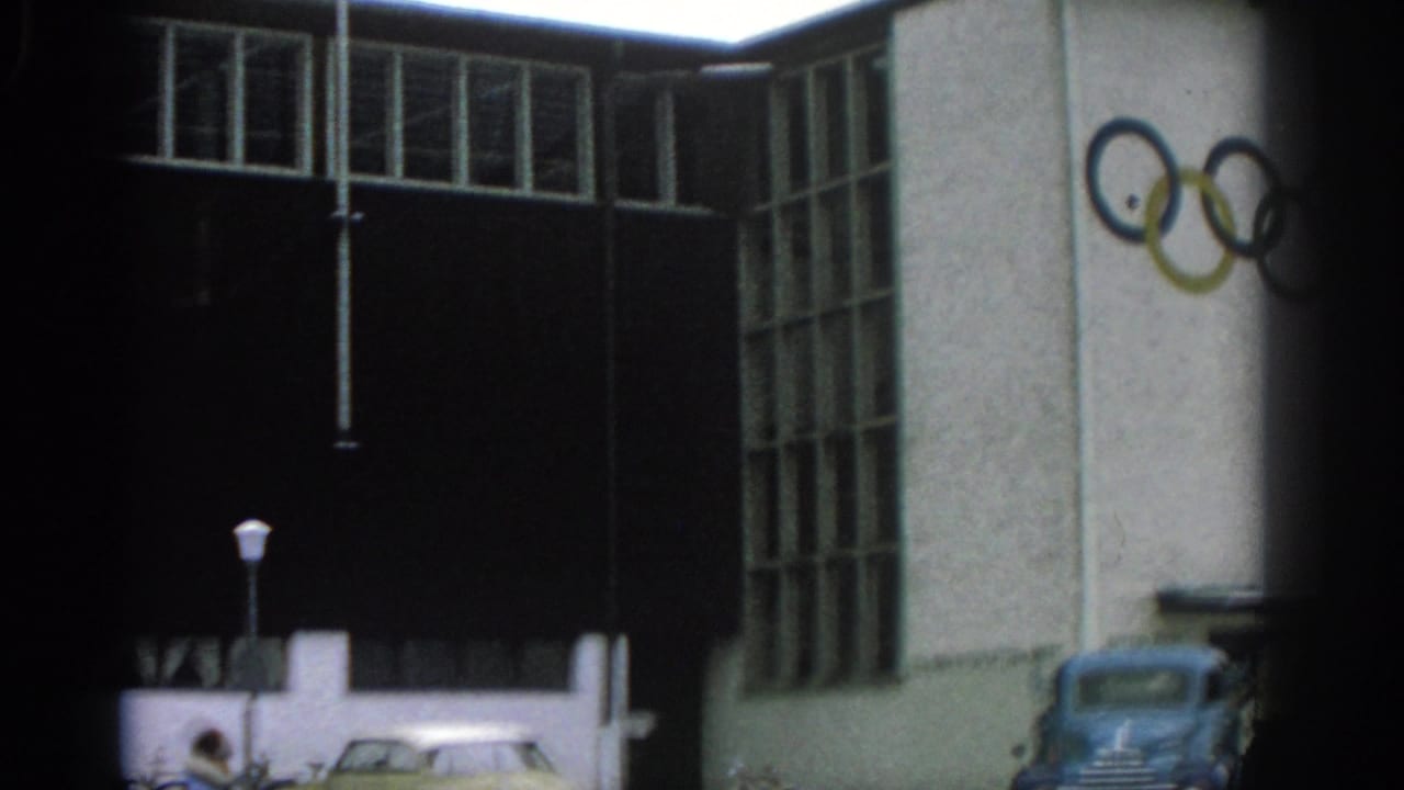 1963 Munich home-movie frame showing a modernist building with Olympic rings on the facade, a blue truck, a light-colored car, and a lamppost near the entrance.
