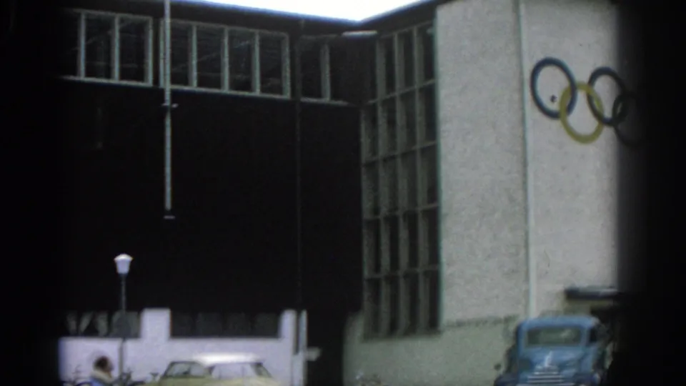 1963 Munich home-movie frame showing a modernist building with Olympic rings on the facade, a blue truck, a light-colored car, and a lamppost near the entrance.