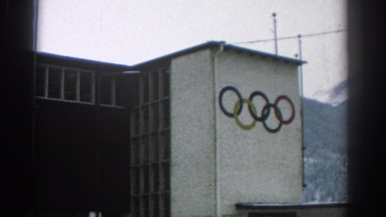 Building corner with Olympic rings, tall windows, roof antennas, and mountains behind.