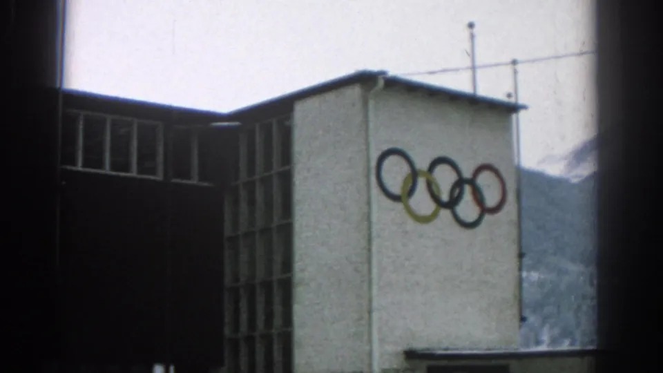 1963 Munich frame of a building corner marked with Olympic rings, with tall window bays, rooftop antennas, and a mountain slope visible in the background.