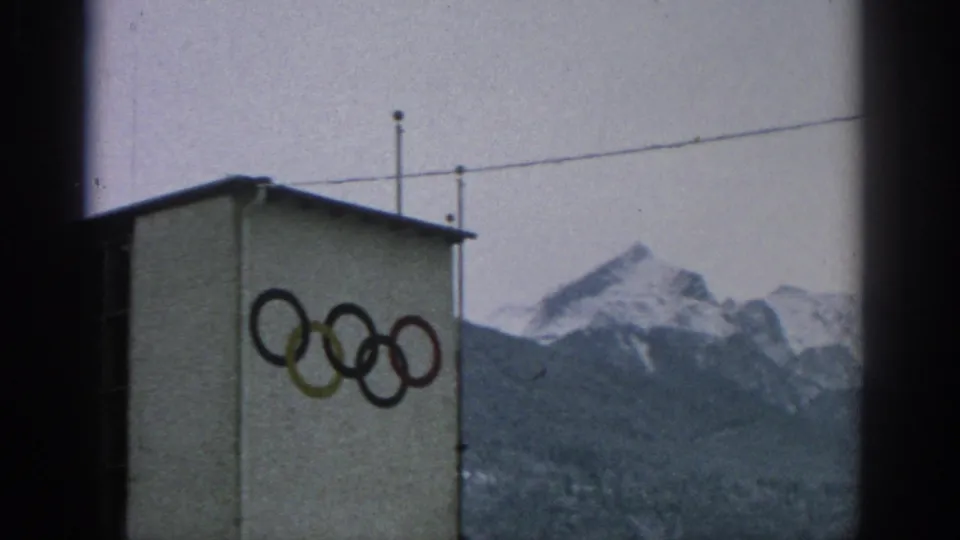 1963 Munich home-movie view of Olympic rings painted on a building wall, with overhead wires and snow-capped alpine peaks beyond.