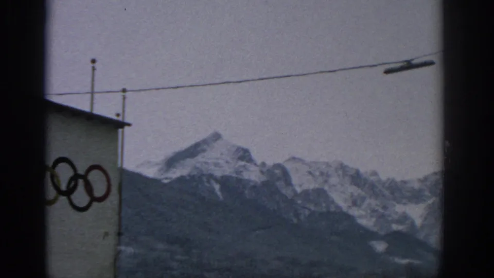 1963 Munich frame with a partial view of the Olympic-ring facade at left, overhead utility wire across the sky, and snow-capped mountains in the background.