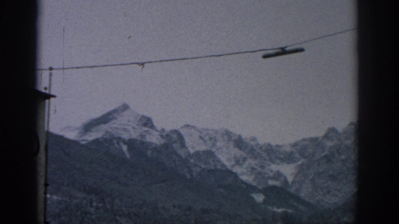 1963 Munich-area home-movie frame showing snow-capped mountains beyond dark window borders and an overhead cable.