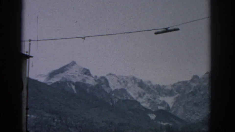 1963 Munich-area home-movie frame showing snow-capped mountains beyond dark window borders and an overhead cable.
