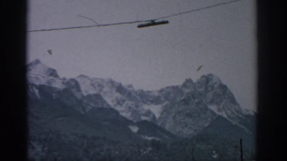 1963 Munich-area alpine view with a cable line above rugged snowy peaks, partly obscured by a dark window frame.