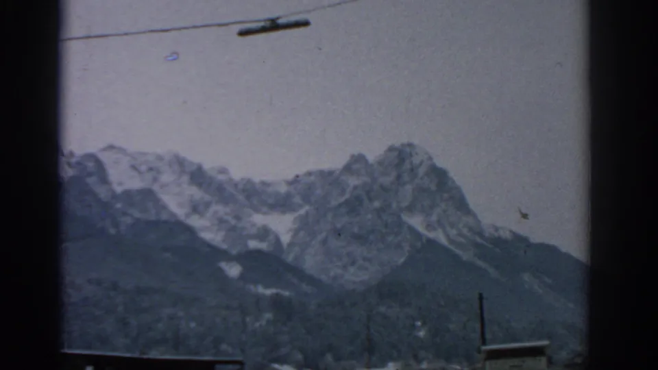 1963 Munich-area frame of jagged snow-capped mountains, overhead gondola cable hardware, and a dark lower hillside.