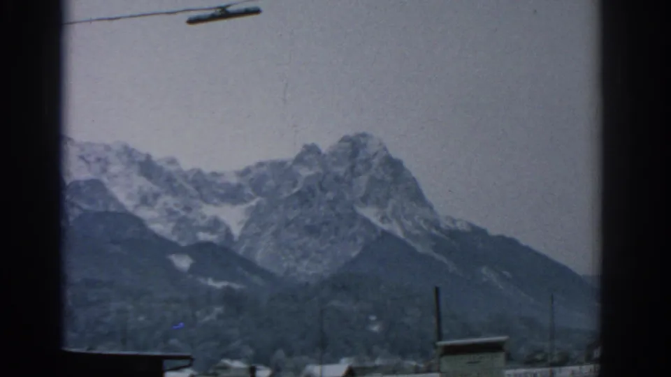 1963 Munich-area home-movie frame showing a snowy alpine ridge beneath a cable line, with a pole and roof edge below.