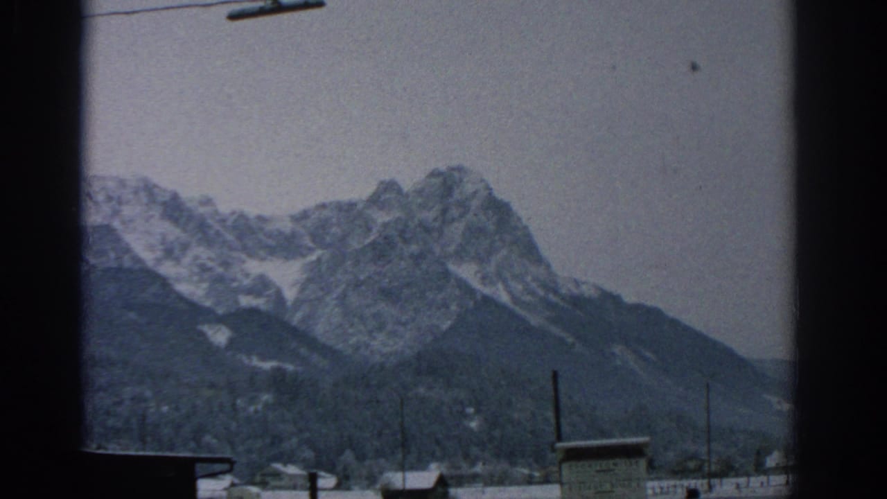 1963 8mm home-movie frame showing snow-dusted alpine peaks, forested slopes, rooftops, and a dark vertical edge suggesting a moving or partially obstructed viewpoint.