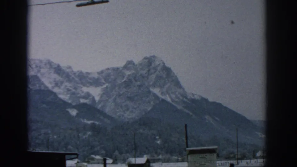1963 8mm home-movie frame showing snow-dusted alpine peaks, forested slopes, rooftops, and a dark vertical edge suggesting a moving or partially obstructed viewpoint.