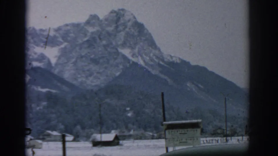 1963 8mm frame of a snow-covered mountain rising behind a small winter settlement, with utility poles and a foreground sign partly visible below the peak.