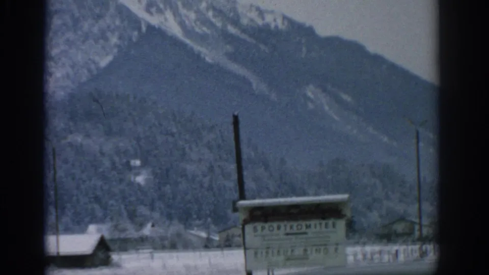 1963 8mm home-movie frame showing a sign that appears to read “SPORTKOMITEE” in front of snowy rooftops, utility poles, and forested alpine slopes.