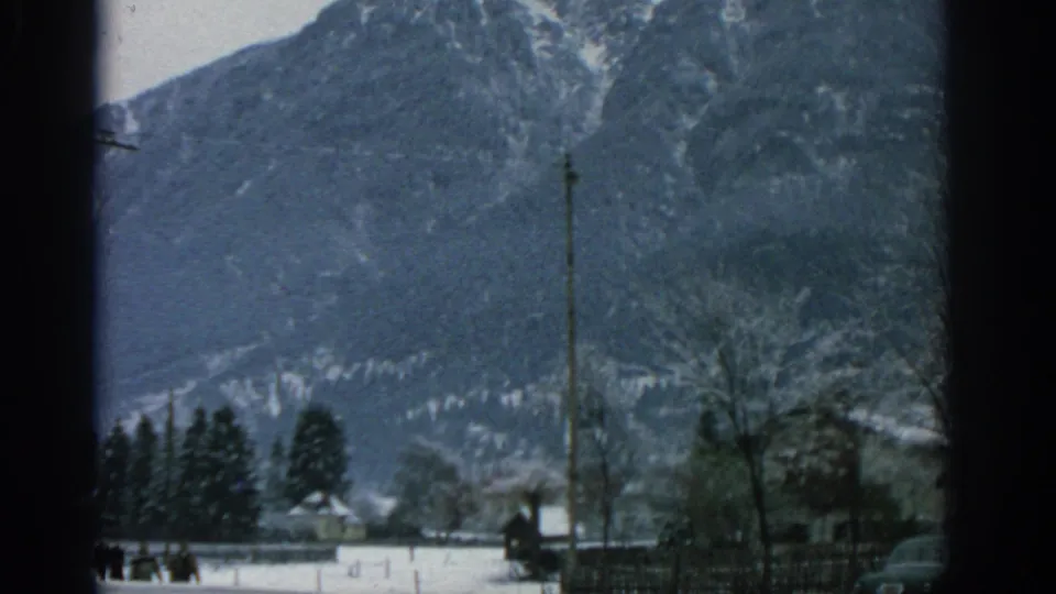1963 8mm frame of a snowy alpine slope behind a village edge, with dark foreground framing, trees, utility poles, small buildings, and a parked car at the lower right.