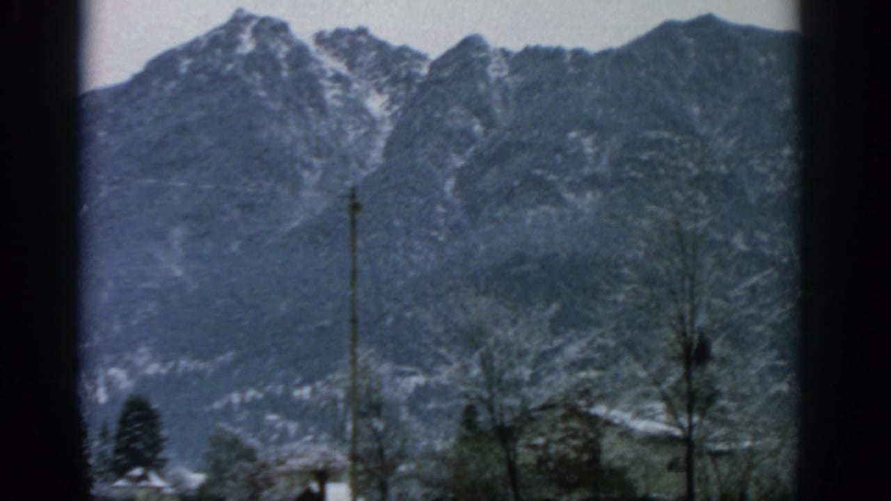 1963 home-movie view of snow-dusted mountain peaks above trees near Munich, with soft 8mm color and dark frame edges.