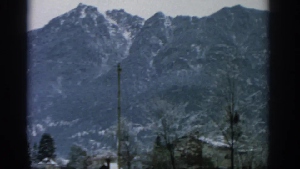 1963 home-movie view of snow-dusted mountain peaks above trees near Munich, with soft 8mm color and dark frame edges.