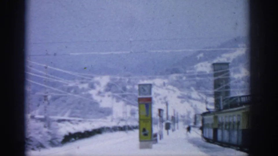 Snow-covered station approach near Munich in 1963, with a clock sign, overhead tram wires, and small figures in the distance.